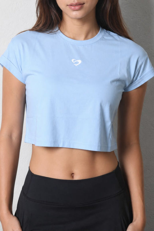Baby Blue Women Crop Tee - SandActive
A woman wearing a light blue cropped short-sleeve athletic T-shirt with a small white heart logo on the chest, paired with black high-waisted leggings, standing against a textured gray wall.