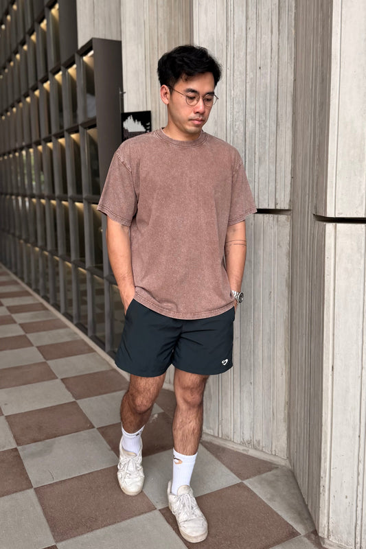 Man wearing a coffee vintage oversized t-shirt and forest green everyday shorts standing in a hallway with a checkered floor.