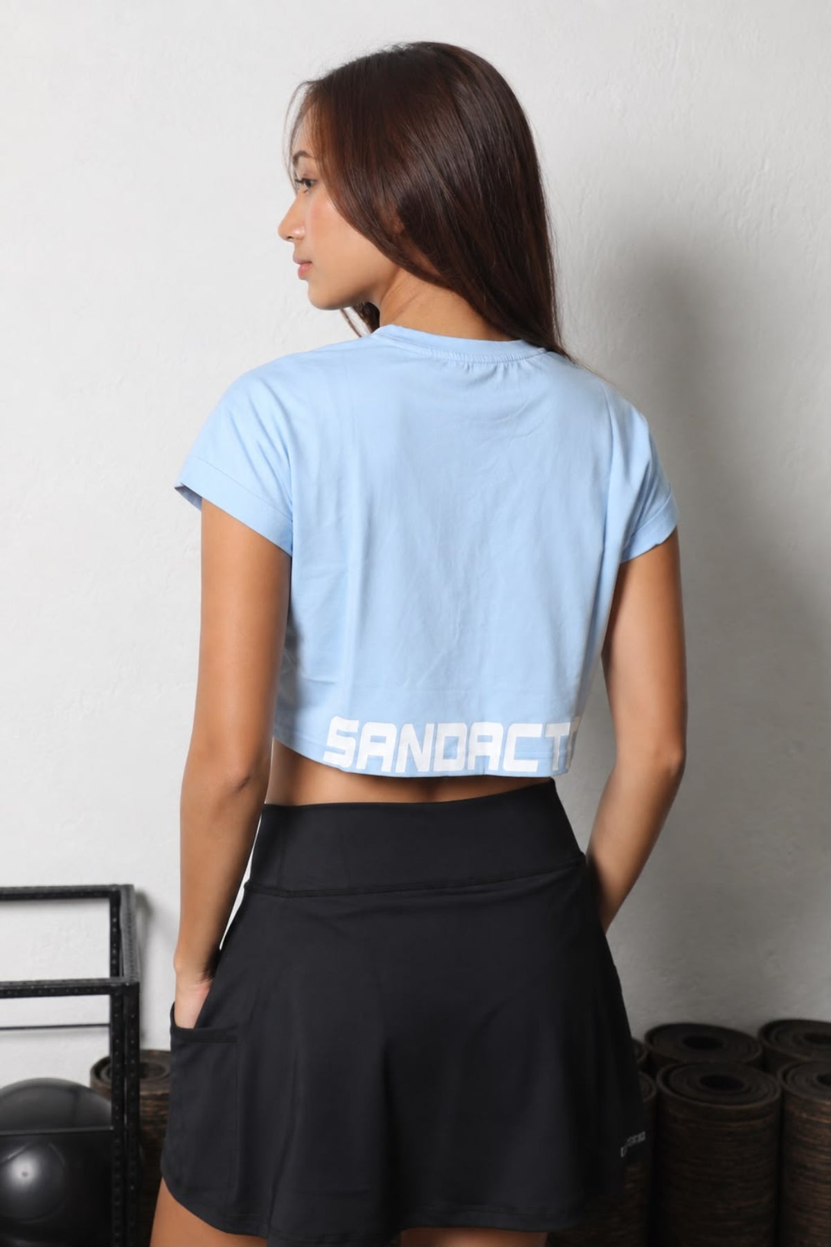 Baby_Blue_Women_Crop_Tee__SandActive 
A woman shown from the back wearing a light blue cropped t-shirt with 'SANACTIVE' printed in white across the lower back, paired with black athletic shorts, set against a plain indoor wall.
