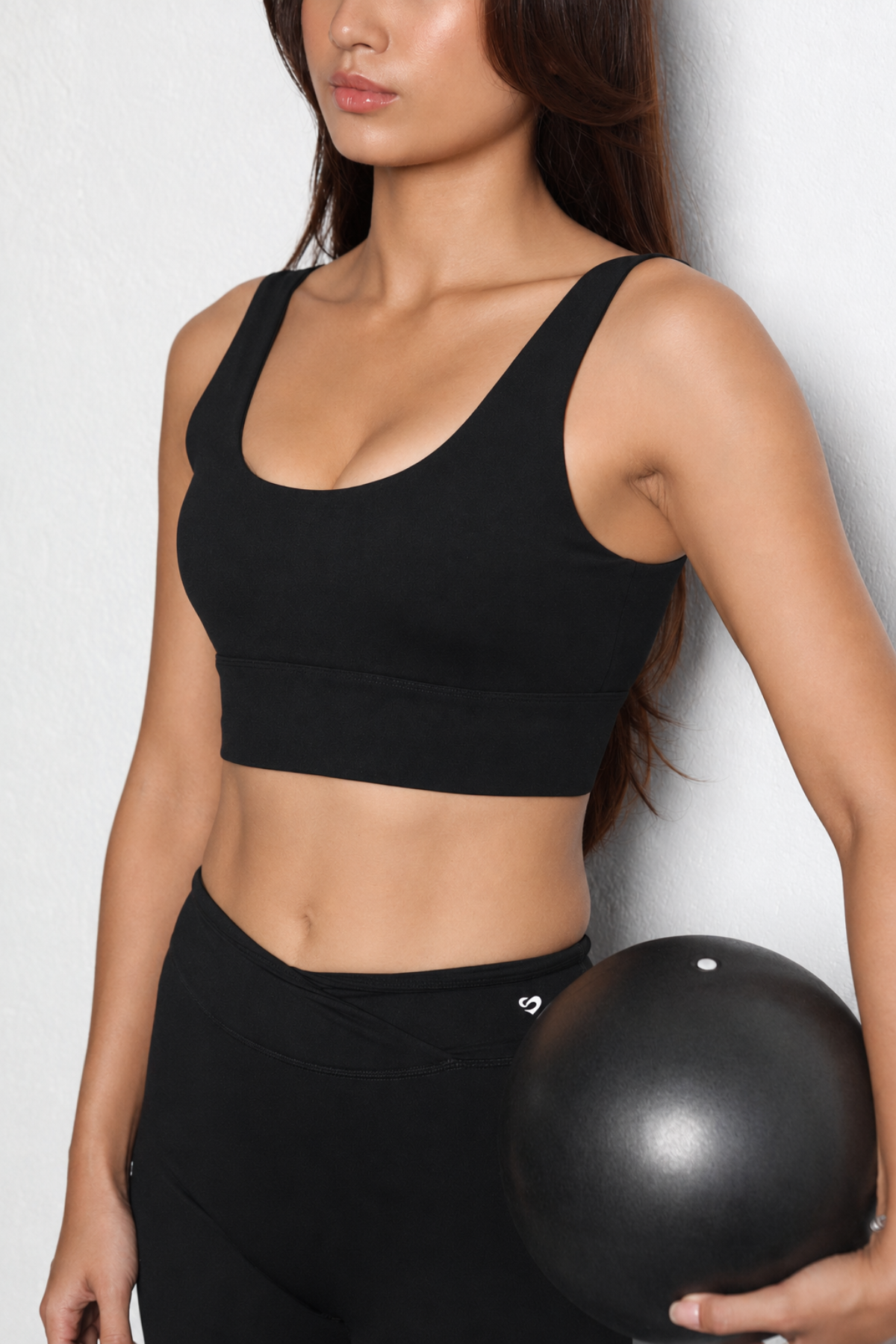 A woman wearing a black scoop-neck sports bra and black leggings, holding a black exercise ball against a light gray wall.