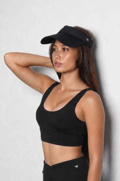 A woman posing in a black athletic sports bra, black leggings, and a black visor against a light gray textured wall.