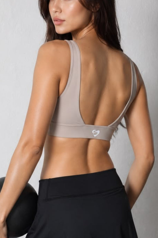 A woman posing from behind in a gym, wearing a beige open-back sports bra and black athletic bottoms while holding a black medicine ball.