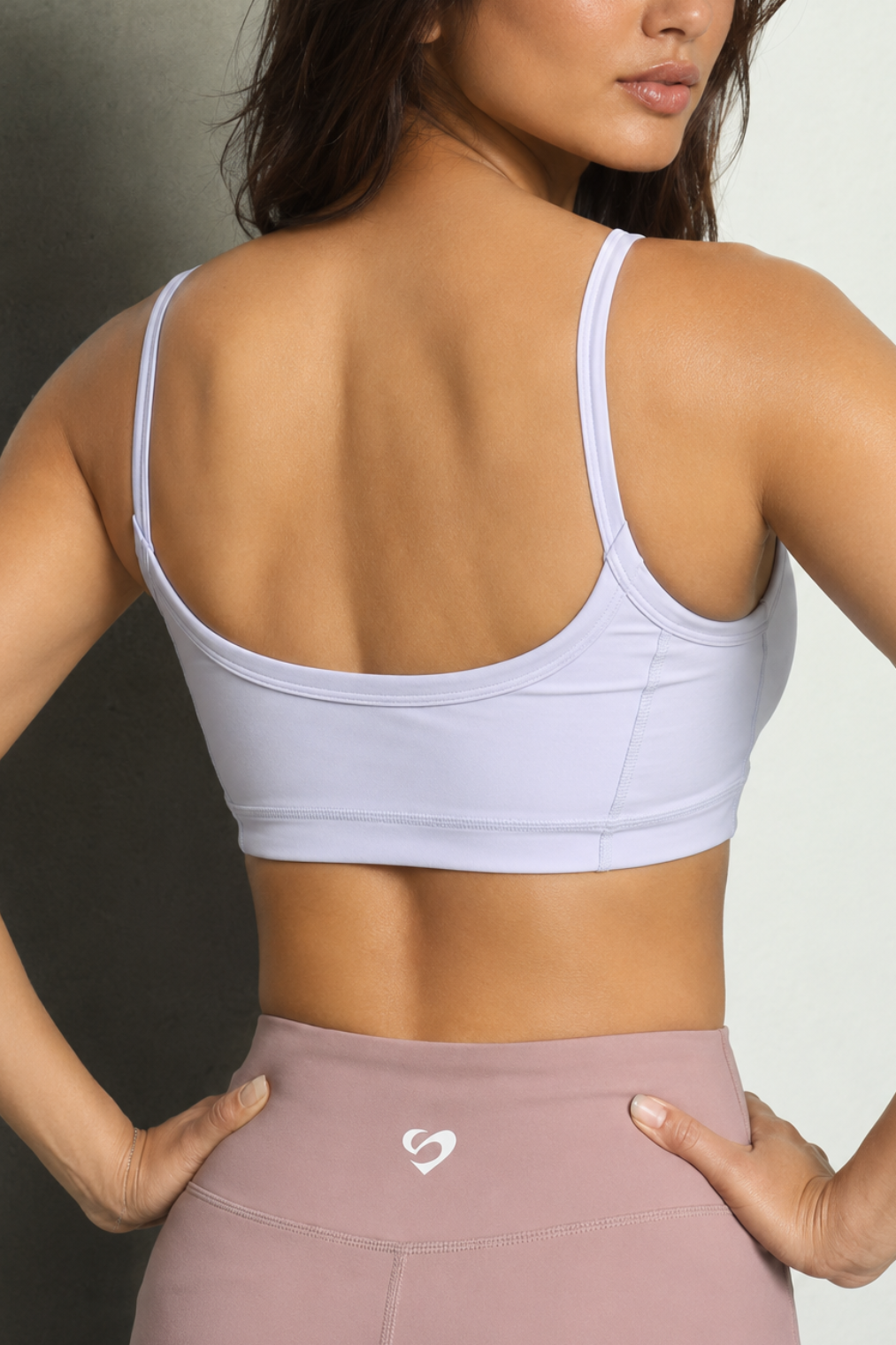 Medium to High Support & Fit 
A woman shown from the back wearing a light lavender sports bra and dusty rose high-waisted leggings with a small white heart logo on the waistband, set against a neutral studio background.