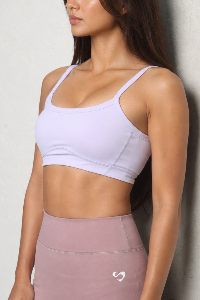 Medium to High Support & Fit 
A woman wearing a light lavender sports bra and dusty rose high-waisted leggings with a small white logo, posing against a textured concrete wall.