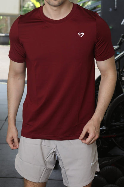 Man wearing maroon everyday t-shirt and ivory white curved hem 8" shorts standing indoors with a bike and equipment in the background - Sandactive