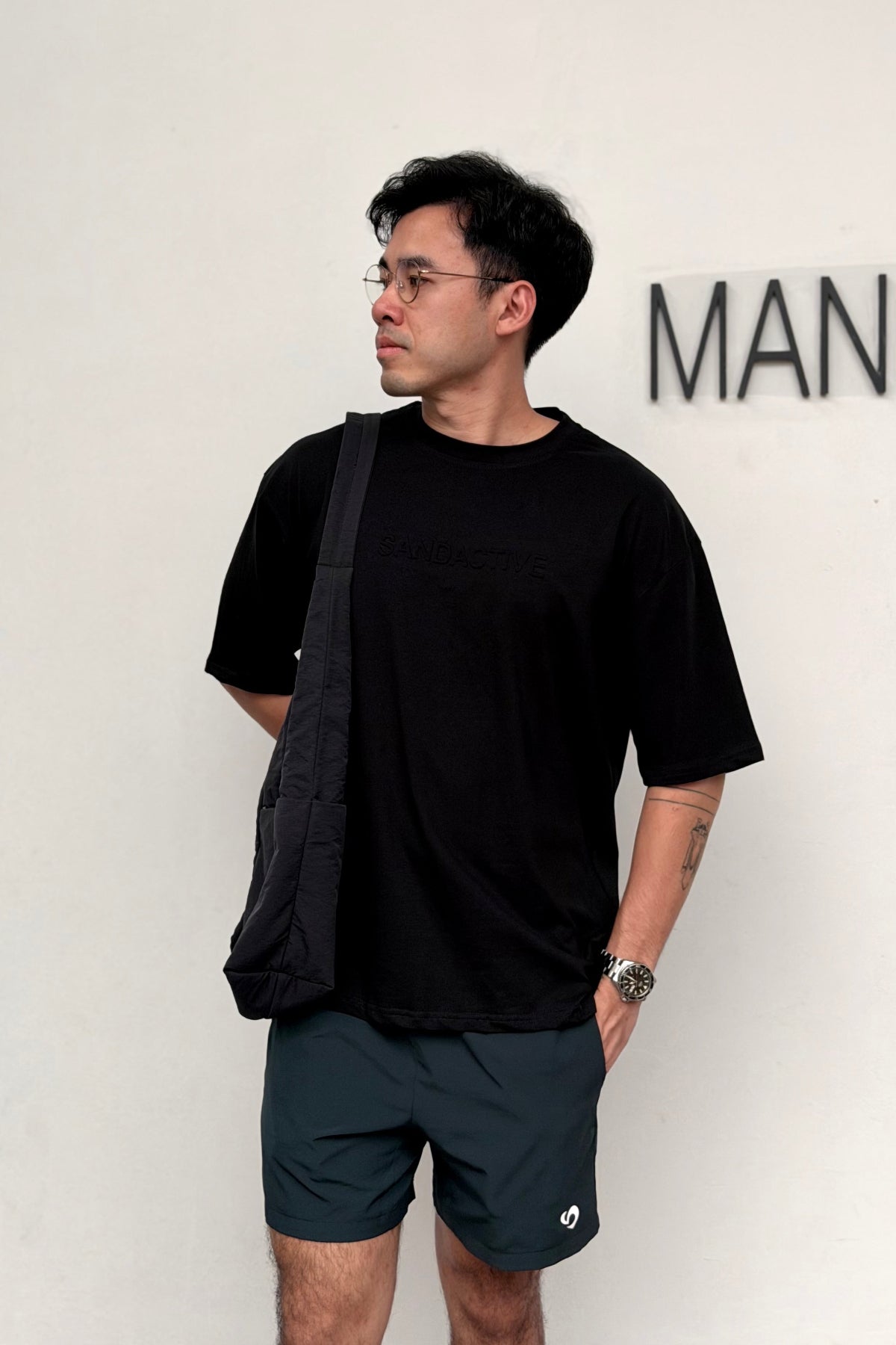 Man wearing a black t-shirt and dark shorts with a crossbody bag, standing against a white wall.