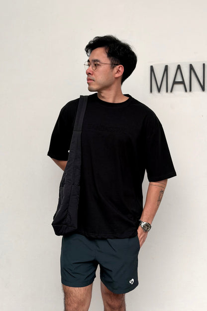 Man wearing a black t-shirt and dark shorts with a crossbody bag, standing against a white wall.