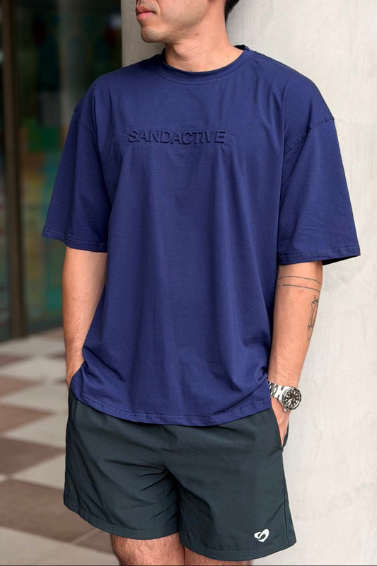 Man wearing a navy blue t-shirt with embossed SandActive name and forest green everyday shorts, standing against a blurred indoor background.