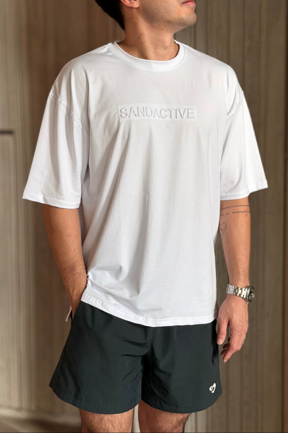 Man wearing a white t-shirt with embossed SandActive brand and forest green everyday shorts standing indoors.