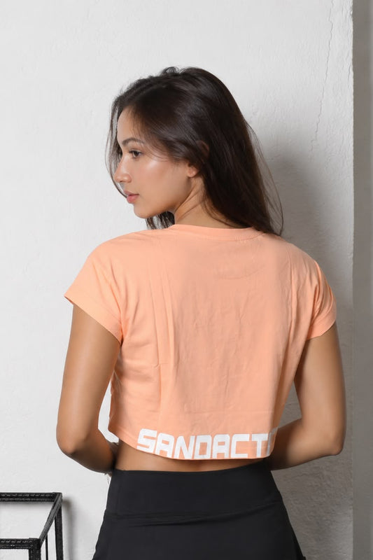 Women's Crop Tee - Orange Crop Tee - SandActive 
A woman wearing a orange-colored crop top with 'SANDACTIVE' printed in white across the back, paired with black bottoms, standing against a white wall.