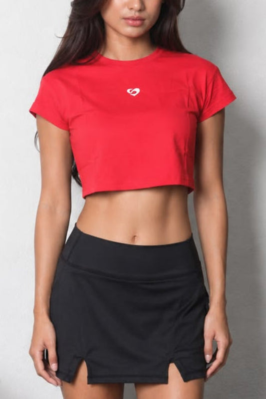 Women's Red Crop Tee - Chic & Comfortable Wear - SandActive 
A woman stands barefoot in a gym setting, wearing a bright red short-sleeve crop top with a small white heart logo on the chest and a black athletic mini skirt with side slits.